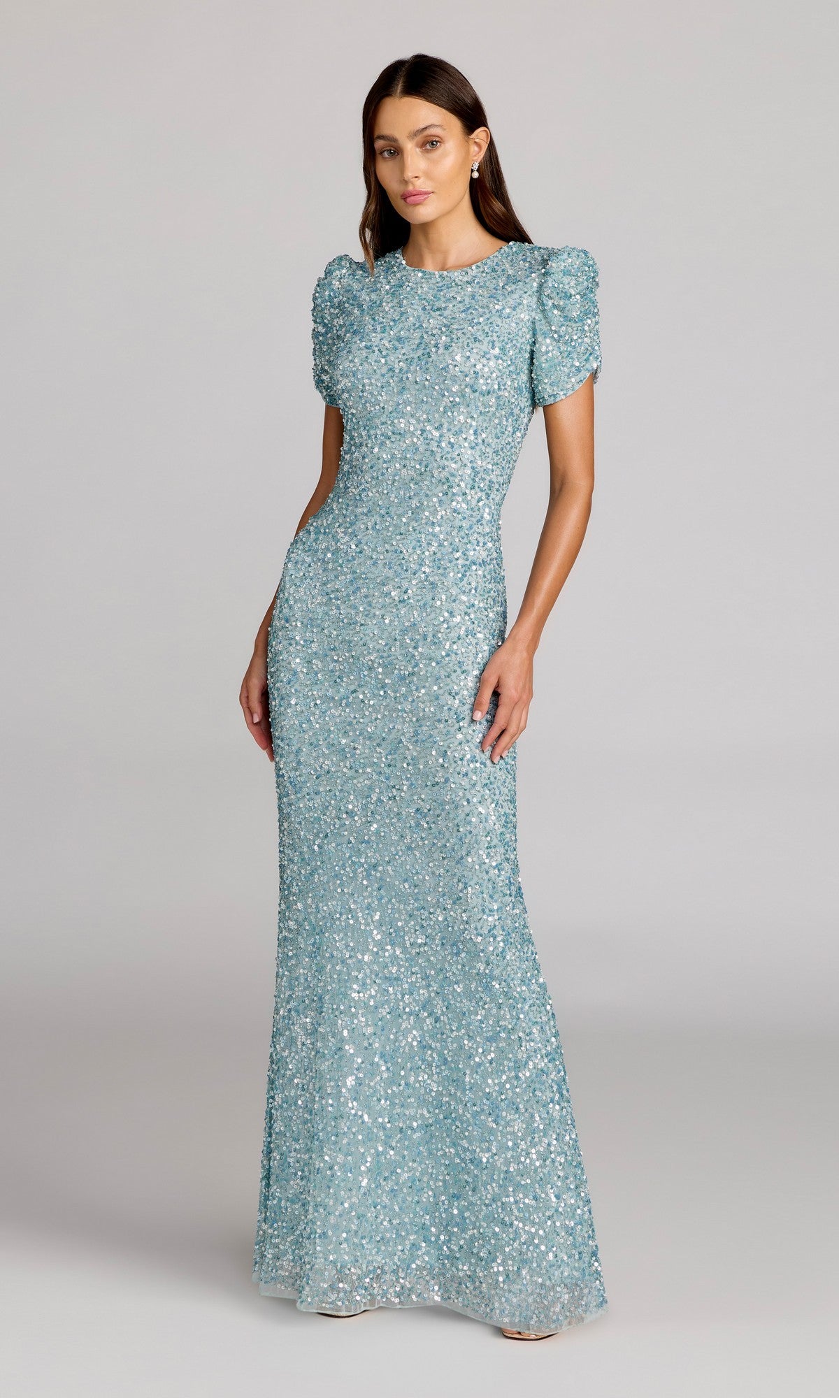 A woman stands against a plain background wearing the Mac Duggal 24003, a light blue, floor-length sequin formal dress with short puff sleeves.